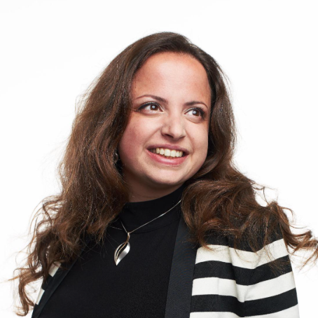 A woman with long, wavy brown hair, smiling and looking to the side, wearing a black top, a black and white striped blazer, and a silver necklace with a pendant, against a white background.