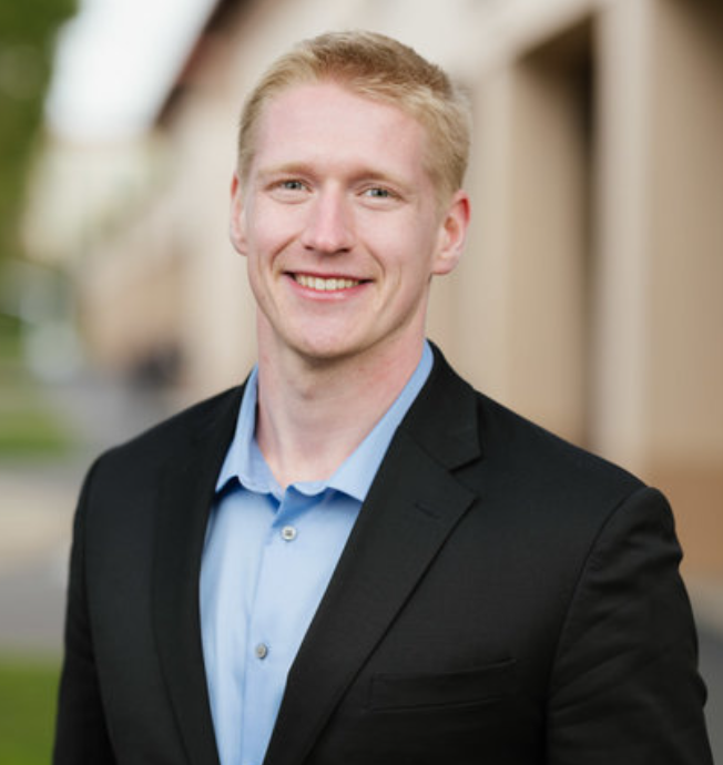 A smiling young man with short blond hair, wearing a dark suit jacket and a light blue collared shirt, standing outdoors with blurred background.