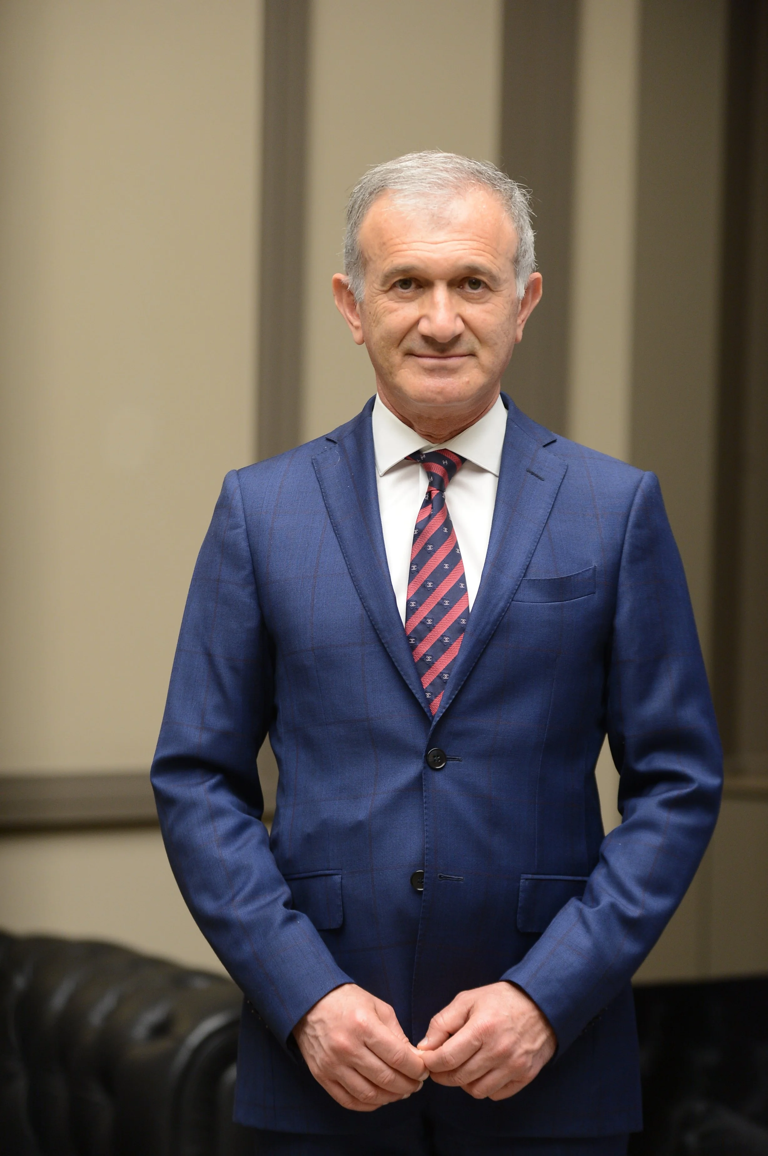 A middle-aged man in a blue suit, white shirt, and red patterned tie standing indoors against a blurred background. He has short gray hair and a slight smile, with his hands clasped in front of him.