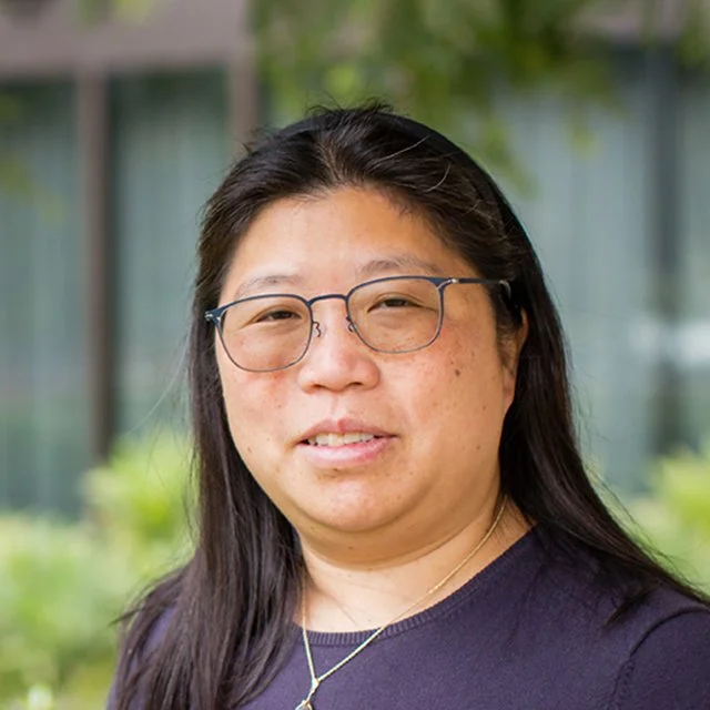 A woman with long black hair, glasses, and a necklace, standing outdoors with a blurred background of greenery and a building.