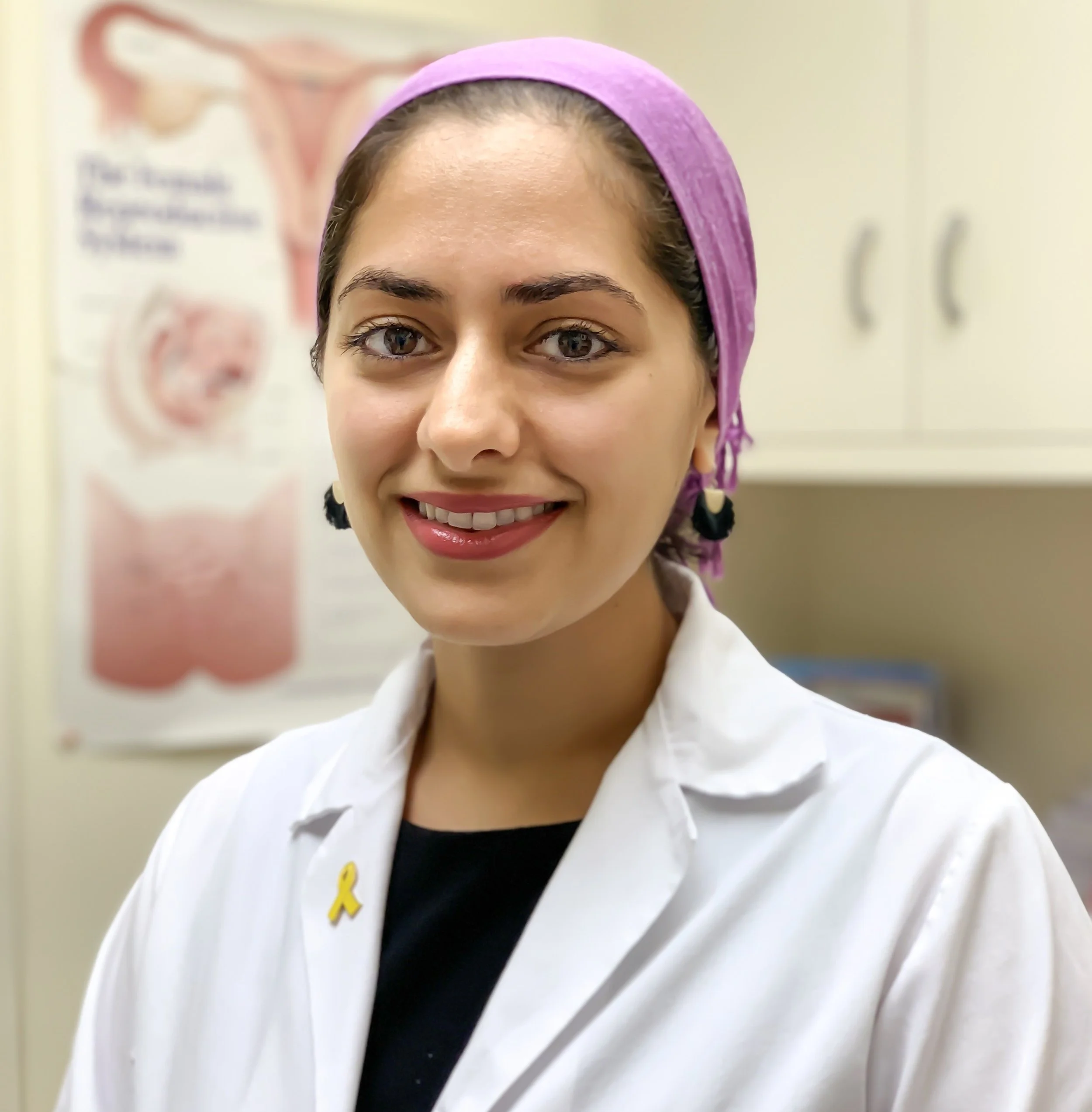 Close-up of a woman with dark hair, wearing a purple headscarf and white blouse, smiling in front of a medical poster about the female reproductive system.