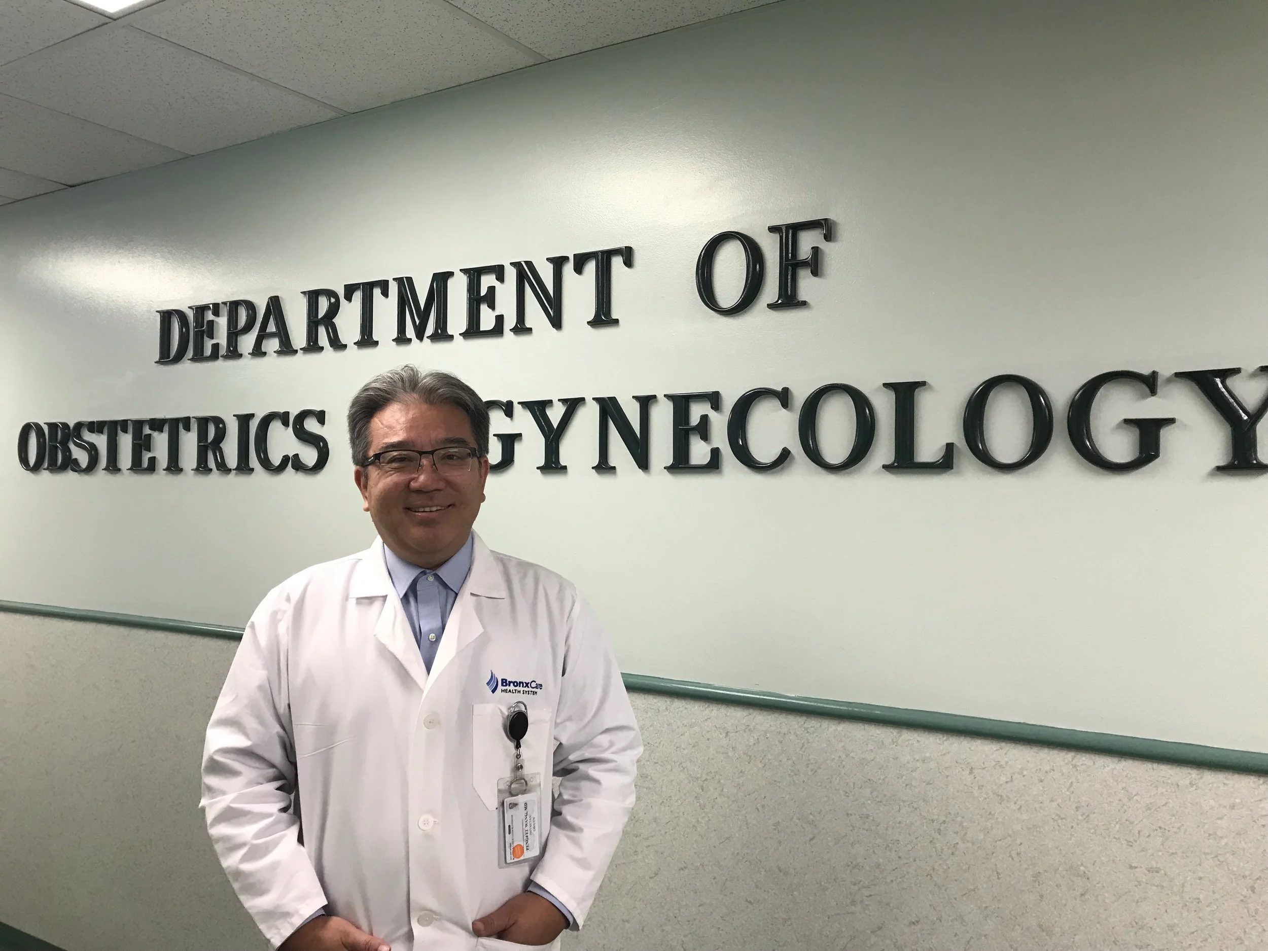 A man in a white medical coat smiling at the camera, standing in front of a sign that reads 'Department of Obstetrics & Gynecology.'