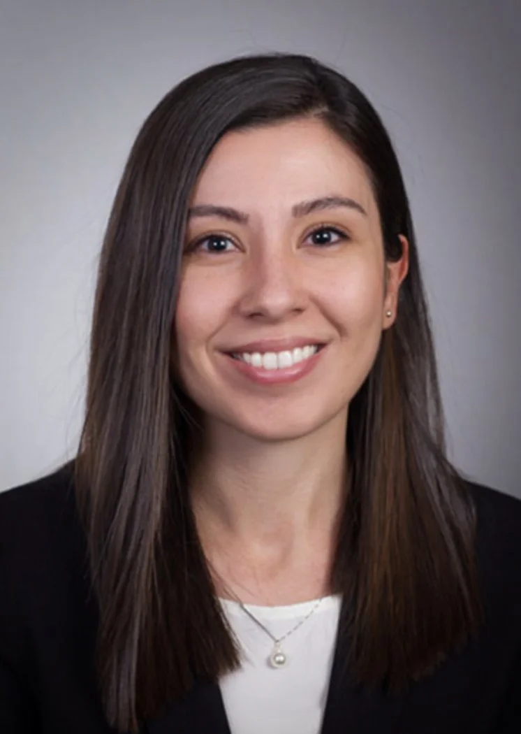 A young woman with straight brown hair and light skin, smiling, wearing a black blazer, white top, pearl necklace, and small earrings, against a gray background.