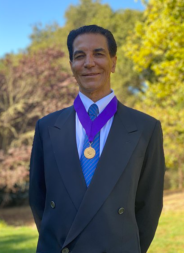 A man in a suit wearing a gold medal around his neck outdoors with trees and blue sky in the background.