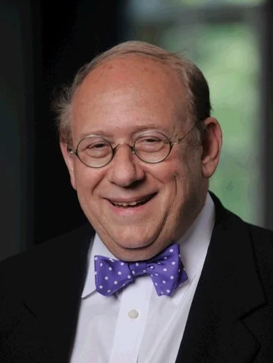 A smiling man with glasses, wearing a black suit, white shirt, and purple polka dot bow tie, in an indoor setting with a blurred window in the background.