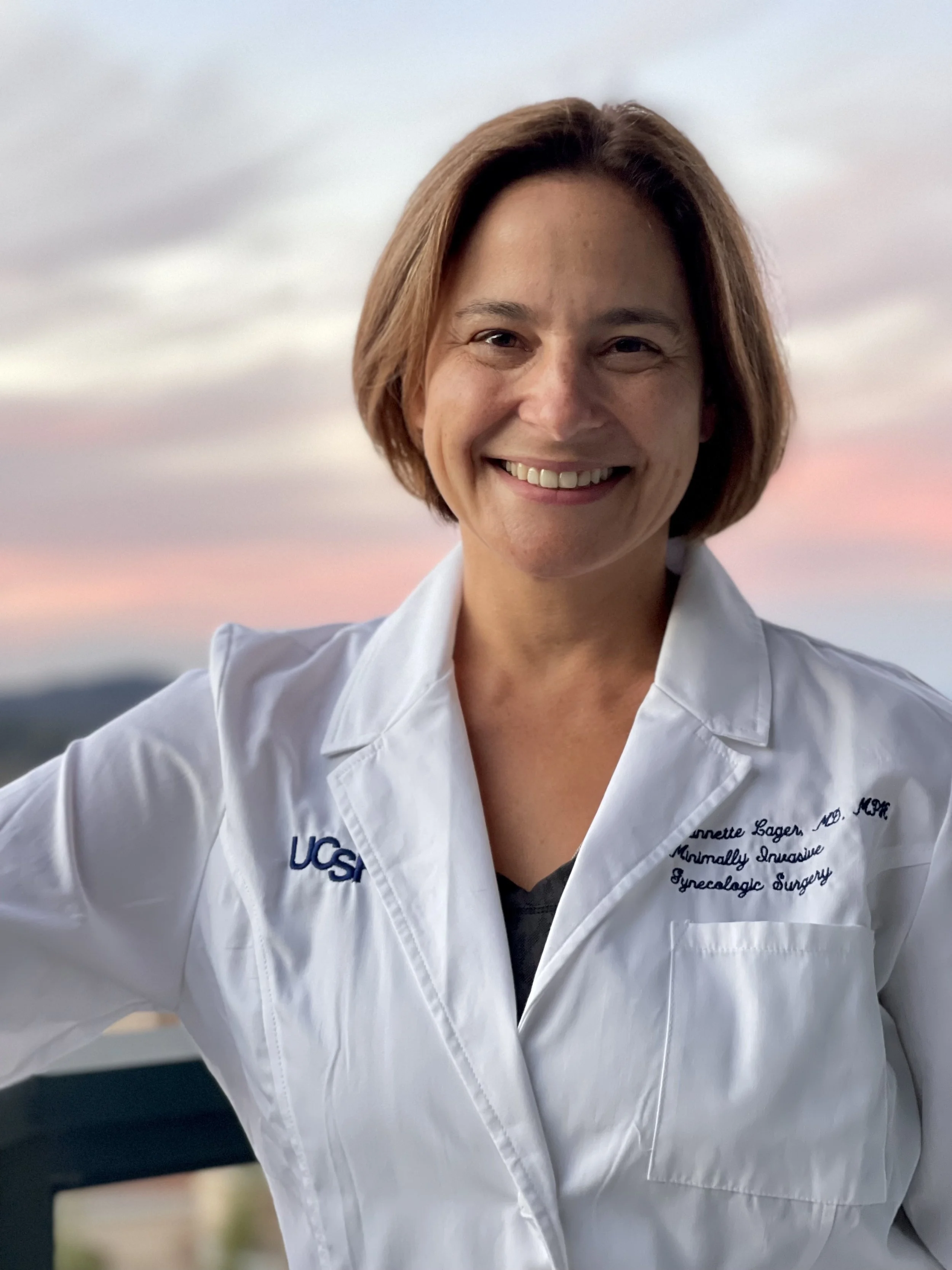 A woman smiling outdoors, wearing a white lab coat with the words 'UC San Francisco' and 'Minimally Invasive Gynecologic Surgery' embroidered on it, with a blurred background of a sunset sky.