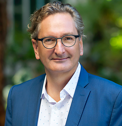 A middle-aged man with gray hair, glasses, wearing a blue suit and white shirt, standing outdoors with greenery in the background.