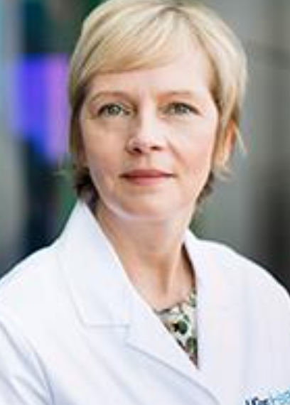 A middle-aged woman with short blonde hair, wearing a white coat, standing in a professional setting.