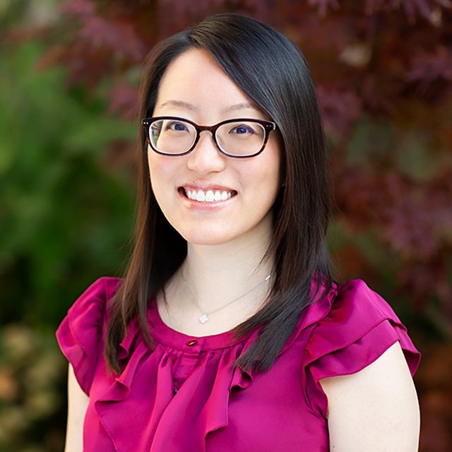 A smiling woman with glasses and long dark hair, wearing a magenta blouse with ruffled sleeves, standing outdoors in front of red and green foliage.