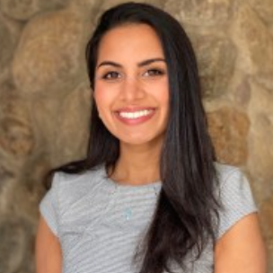 A young woman with long dark hair smiling, wearing a light gray top, standing in front of a stone wall.
