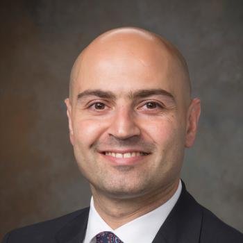 A bald man with a light skin tone wearing a dark suit, white shirt, and patterned tie, smiling against a neutral background.