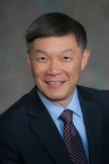 Portrait of a man with short black hair, wearing a navy blazer, light blue shirt, and a purple patterned tie, smiling in front of a gray background.