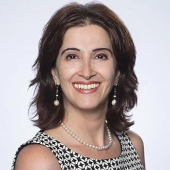 A woman with shoulder-length brown hair, wearing pearl earrings and a necklace, smiling at the camera against a plain background.