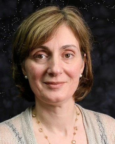 A woman with short light brown hair, wearing pearl earrings, a beige cardigan with gray trim, and a gold necklace, smiling at the camera against a dark, indistinct background.