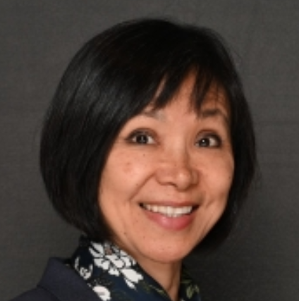 Smiling woman with short black hair wearing a dark blazer and floral shirt against a gray background.
