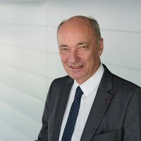 A professional middle-aged man in a dark suit, white shirt, and tie, smiling in a modern office setting.