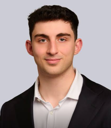 A young man with dark hair and fair skin wearing a white shirt and black blazer, smiling at the camera against a light gray background.