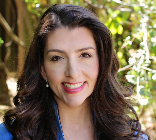 A woman with long dark hair smiling outdoors with sunlight and greenery in the background.