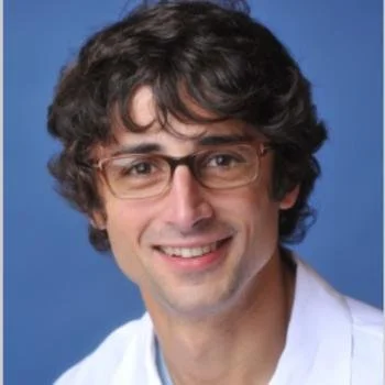 Portrait of a man with curly brown hair, glasses, and a friendly smile, wearing a white coat against a blue background.