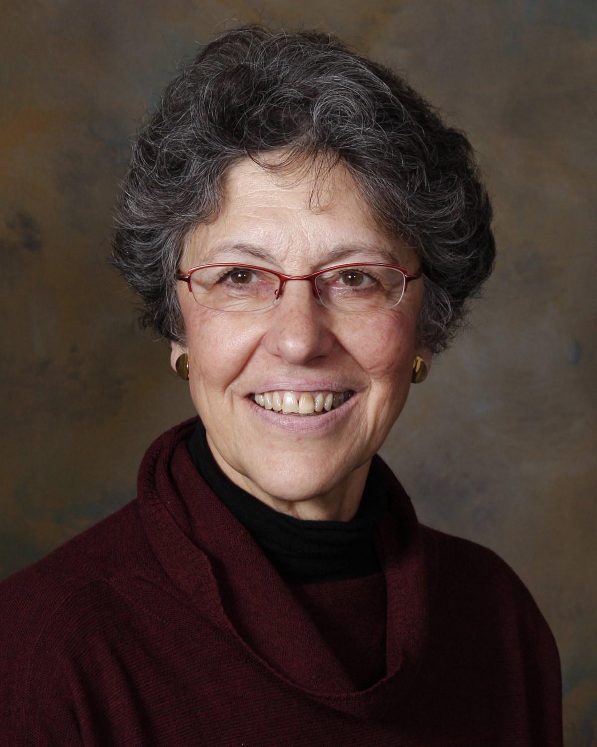 A smiling older woman with short gray hair, glasses, earrings, and wearing a dark red turtleneck sweater standing in front of a brownish background.