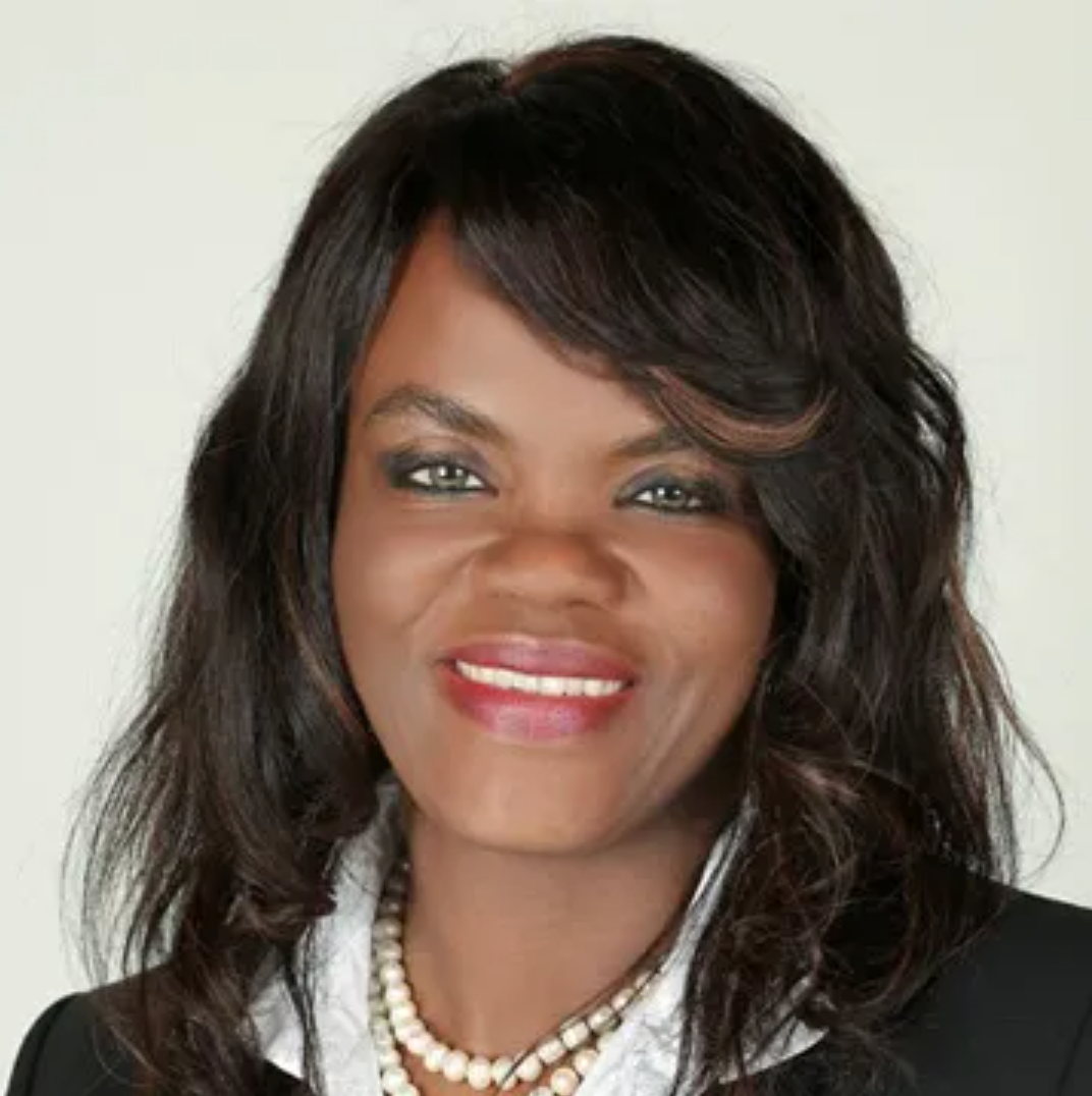 Close-up portrait of an African American woman with shoulder-length wavy hair, wearing a black blazer, white collared shirt, and pearl necklace, smiling against a light background.