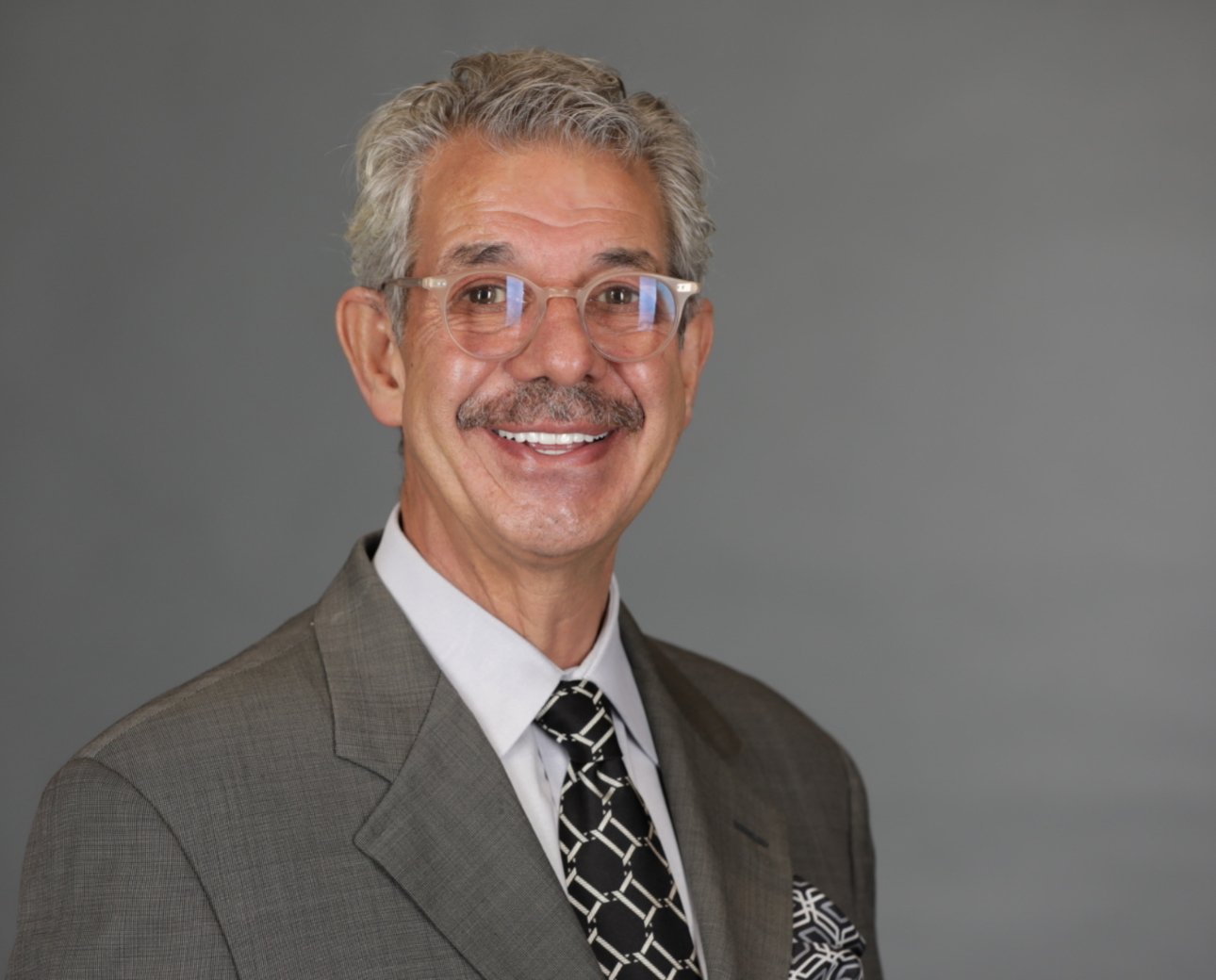 Portrait of a smiling middle-aged man with gray hair, glasses, wearing a suit and tie, against a plain gray background.