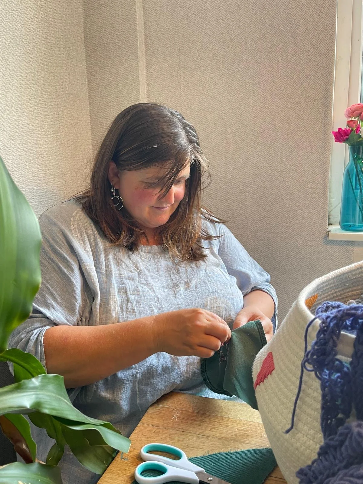 Female artist with dark hair, sat looking down sewing green fabric wearing a blue top, smiling at her work