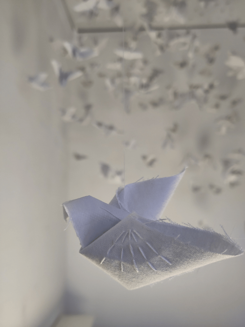Close up photo in the foreground of white origami bird made of cotton shirts, with white stitching on its wing