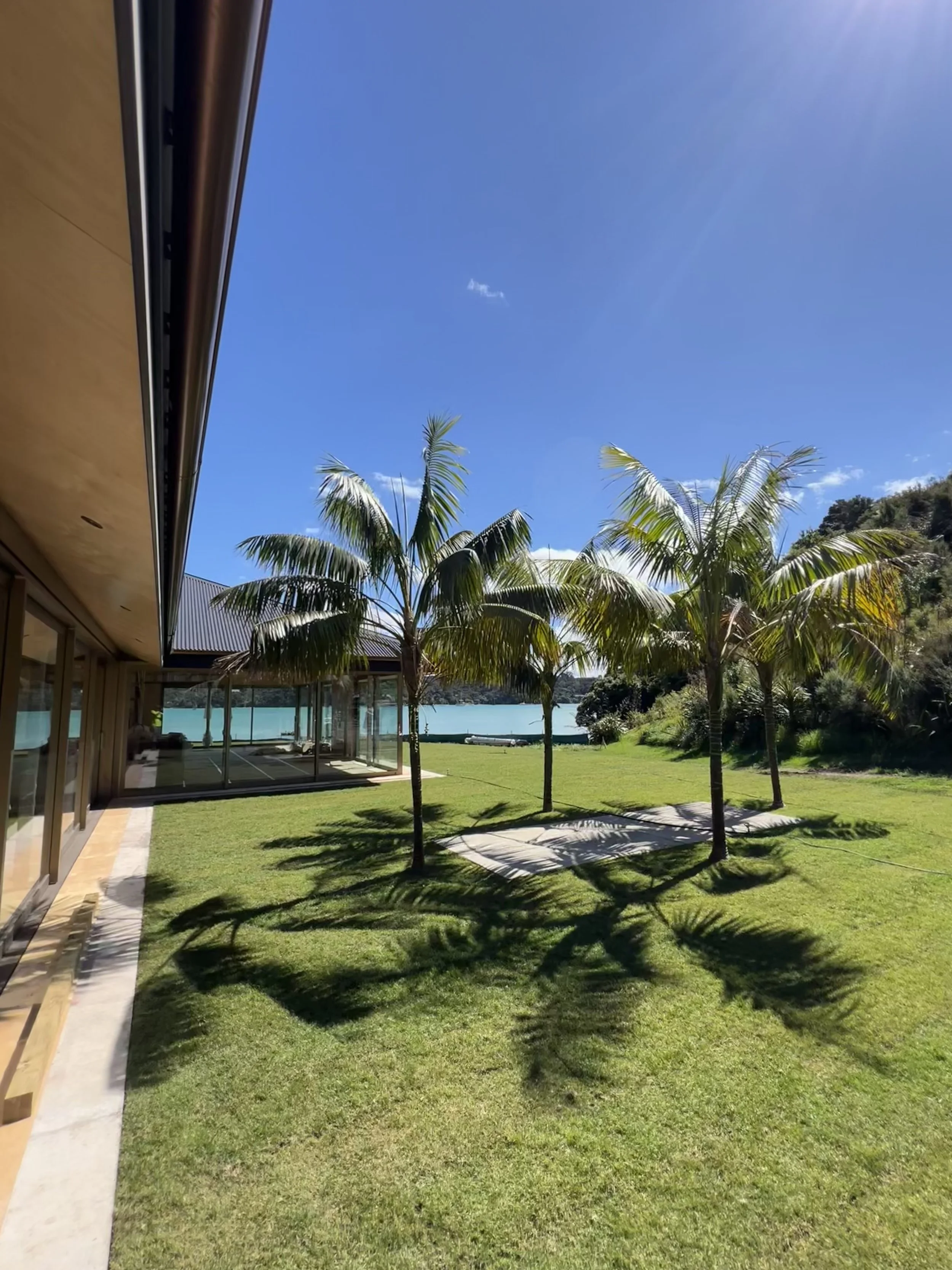 A modern house with a glass wall outdoor patio, lush green grass, and tall palm trees. In the background, there is water and a hill under a bright blue sky with sun rays.