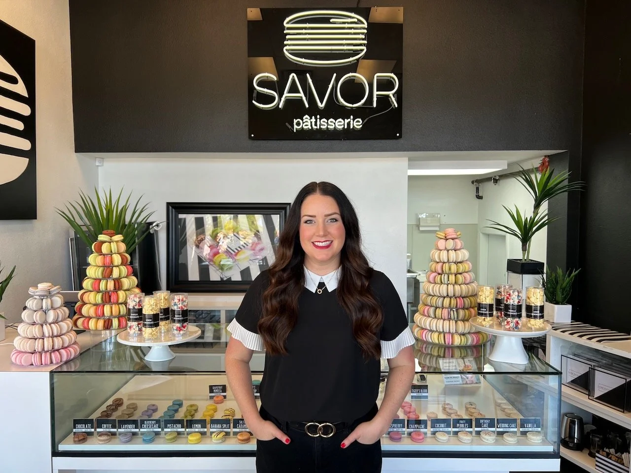 Smiling woman standing in front of a Savor Patisserie display case with colorful macarons and macaron towers.