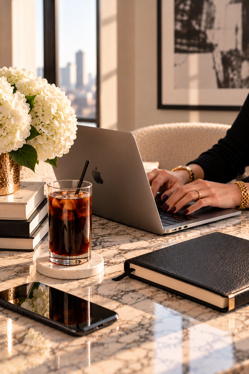 Person working on a laptop at a marble table with white hydrangeas, iced coffee, books, and a city view