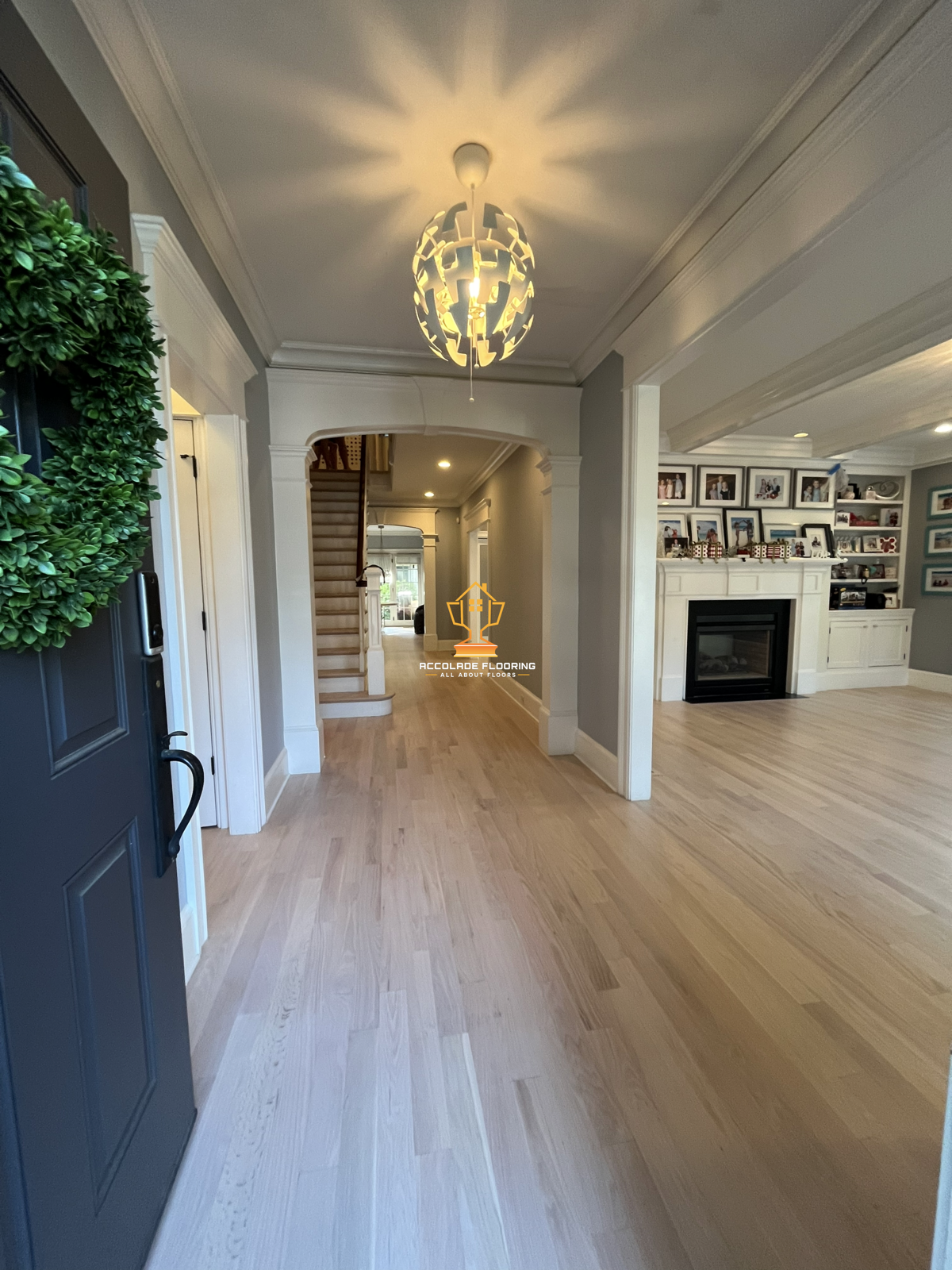 Interior view of a home foyer and living room with light wood flooring, a staircase, a fireplace with a framed family photo display above, and a modern chandelier in the ceiling casting decorative light patterns.