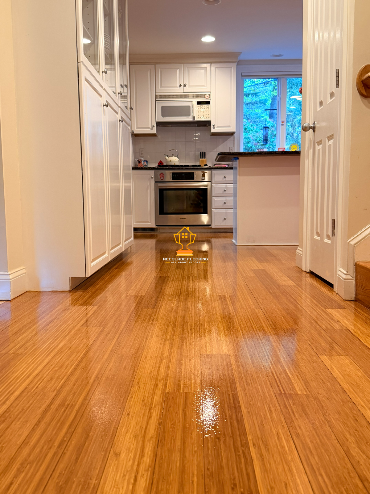 Kitchen with white cabinets, stainless steel oven, microwave, and a window showing blue outside sky and trees, with a polished wooden floor.