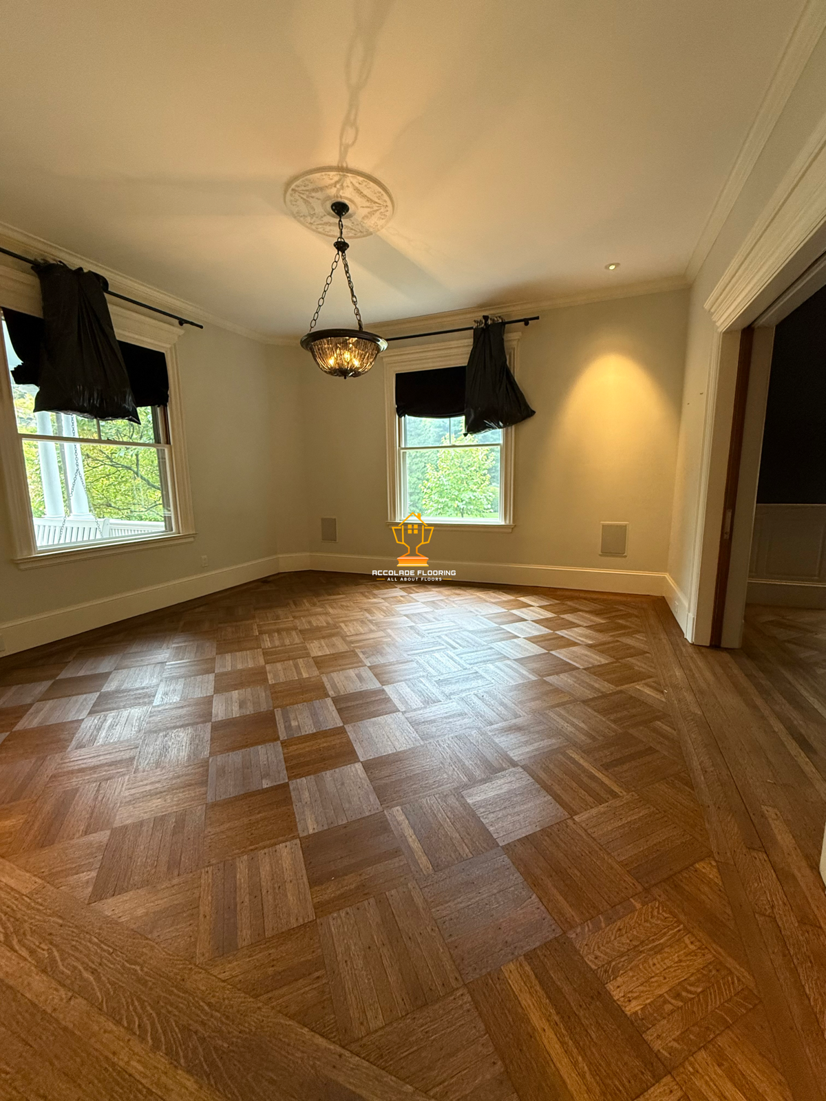 Empty room with hardwood checkered floor, white walls, two windows with black curtains, and a chandelier hanging from the ceiling.