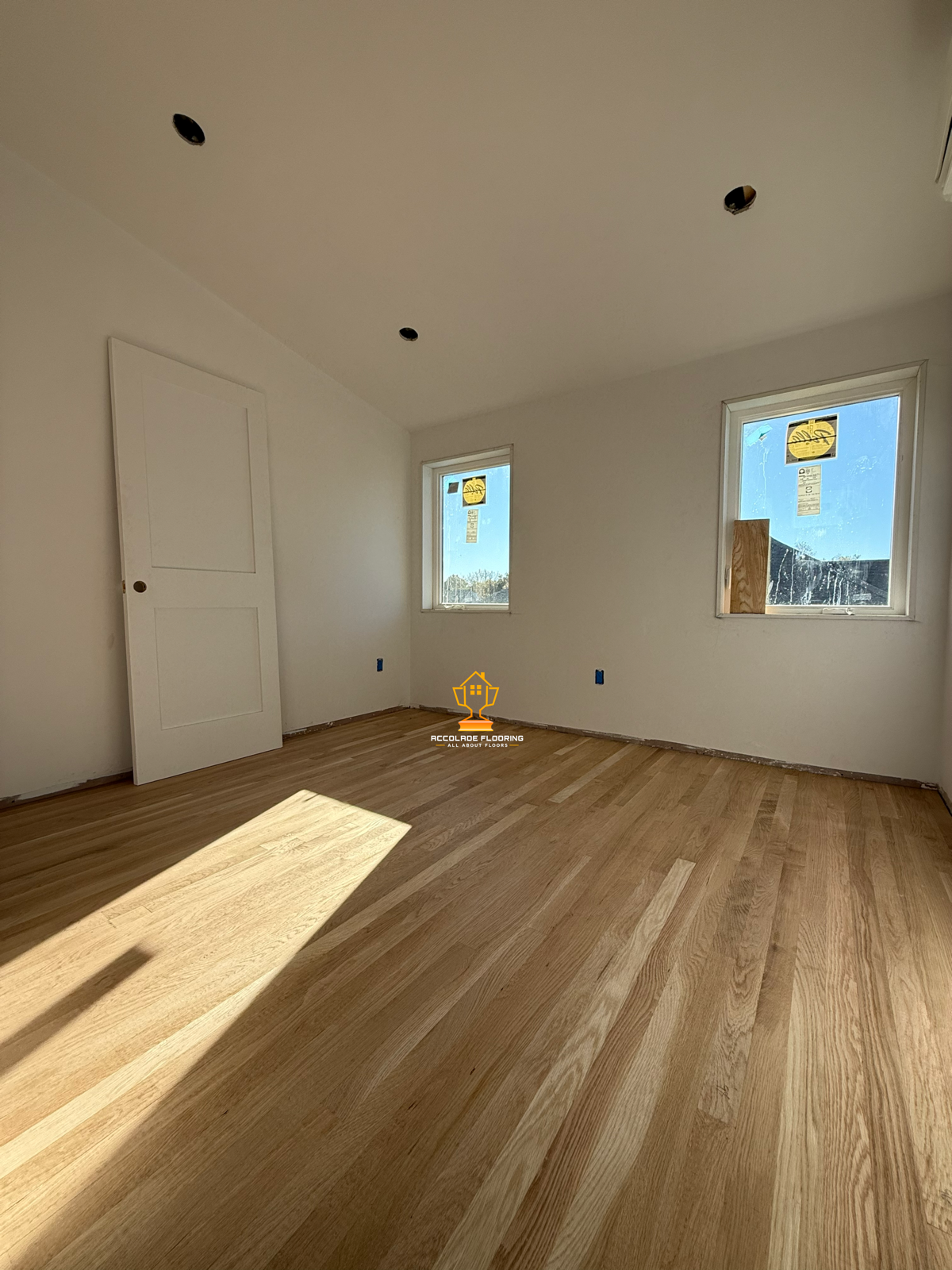 A room under construction with two windows, a door, and new hardwood flooring. The walls are painted white and there are electrical outlets visible. Sunlight is streaming into the room.