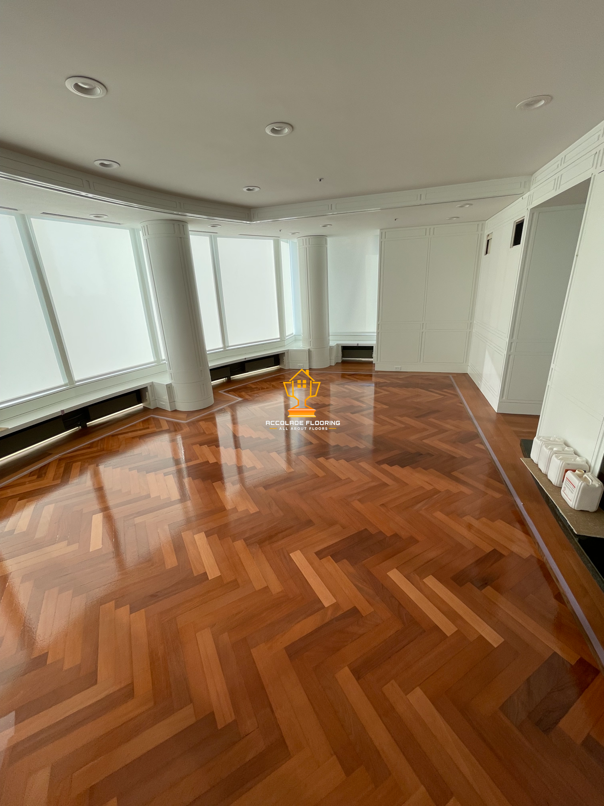 Empty room with large windows, white paneled walls, intricate parquet wood flooring, and ceiling lights.
