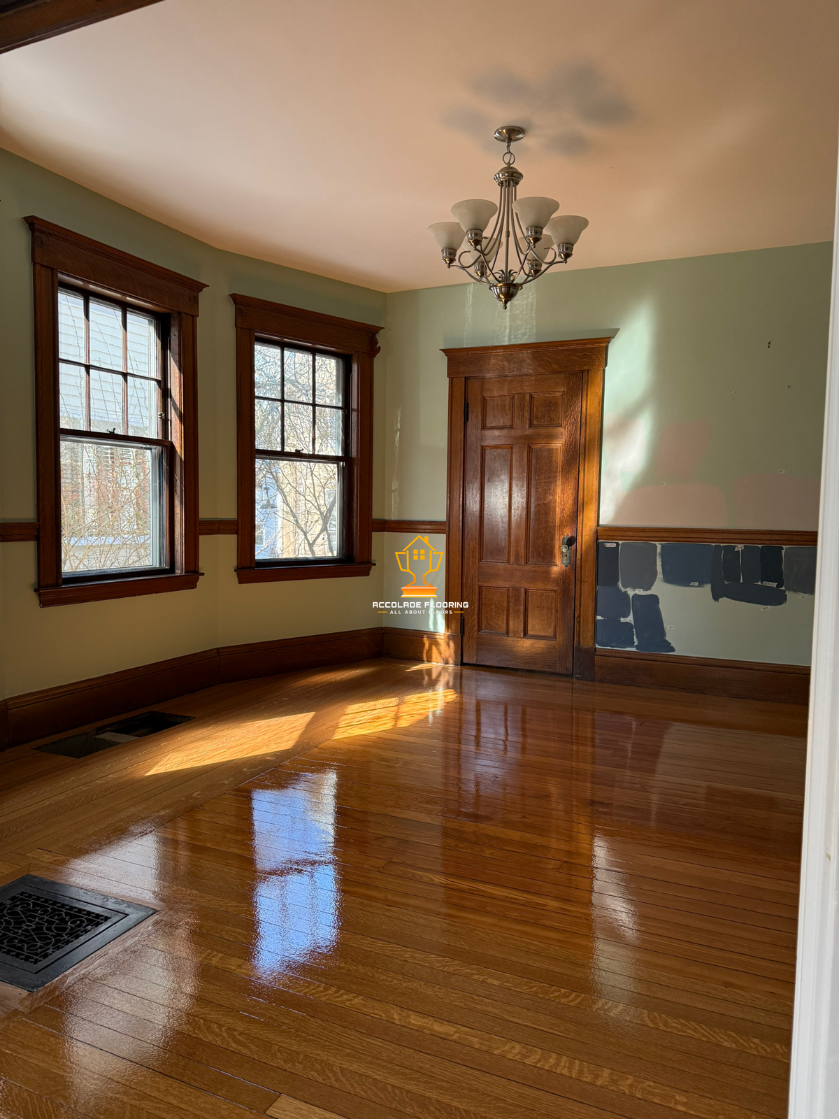 Empty room with hardwood floors, two large windows with wooden trim, a wooden door, and a chandelier hanging from the ceiling. There is a painted patch on the wall near the door.