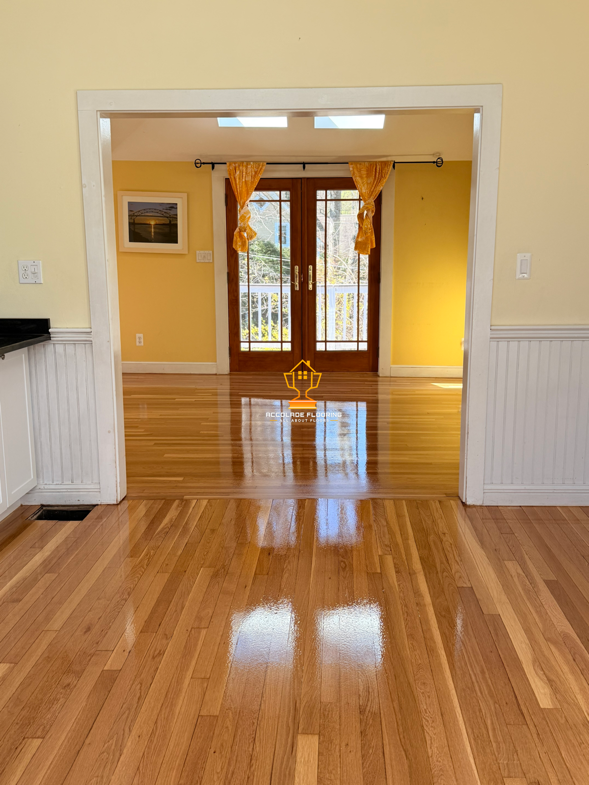 View of a room with polished wooden flooring, yellow walls, and a pair of wooden-framed glass doors leading to a balcony with outdoor scenery. There are orange curtains tied on either side of the doors.