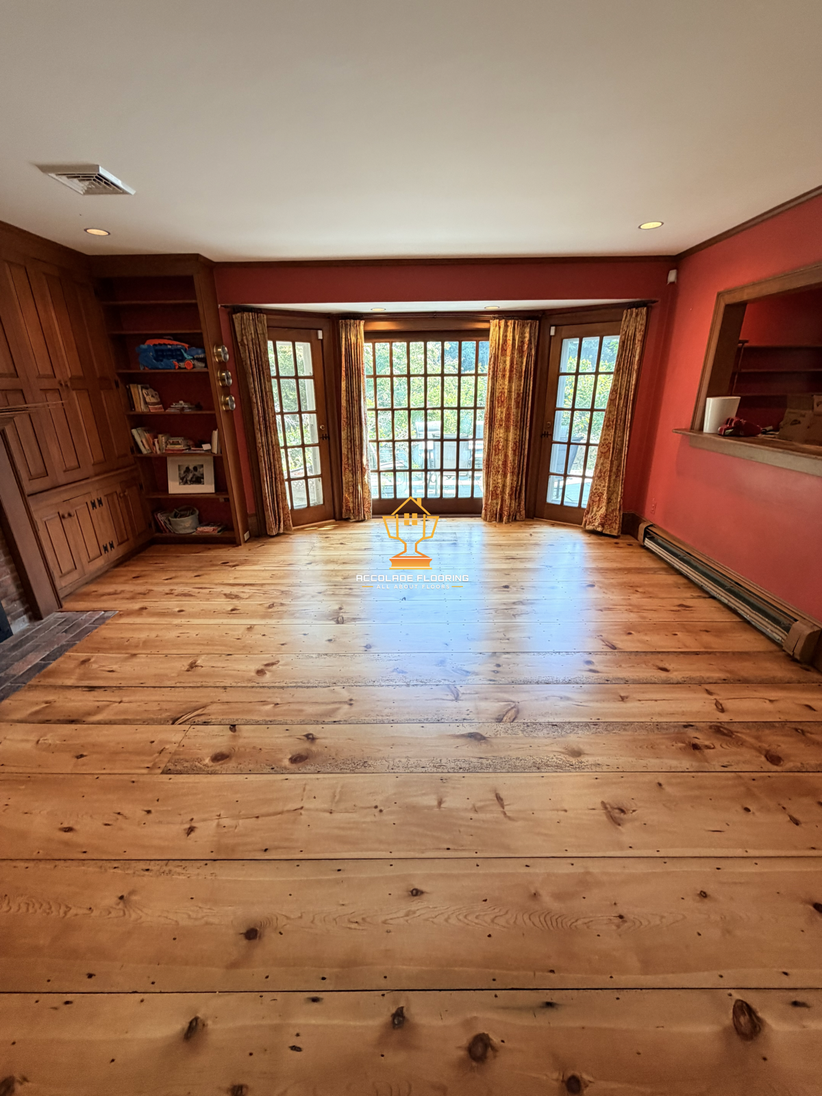 Empty living room with wooden floors, red walls, large glass door with curtains leading to an outdoor deck, built-in wooden shelves and cabinet, and a partial view of a brick fireplace.