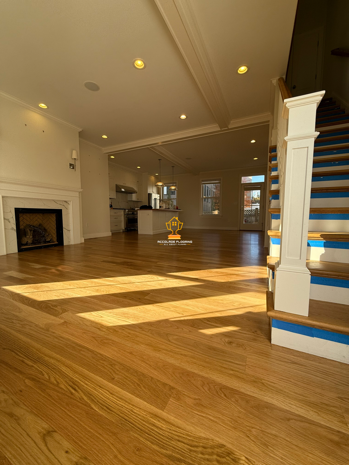 Living room with finished hardwood flooring, white walls, and a fireplace with a marble mantle. The space is bright with natural light coming through windows and a door leading outside. Staircase on the right side has blue painter's tape on the steps