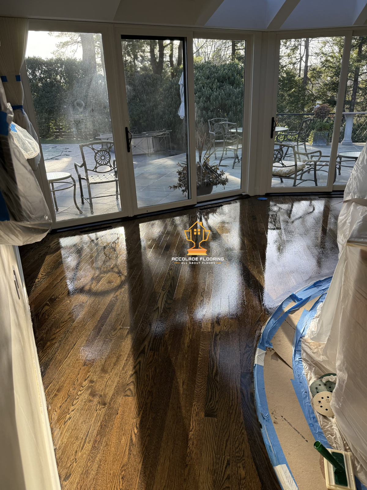 Refinished hardwood floor in a sunlit room with glass sliding doors leading to an outdoor patio with chairs and greenery.