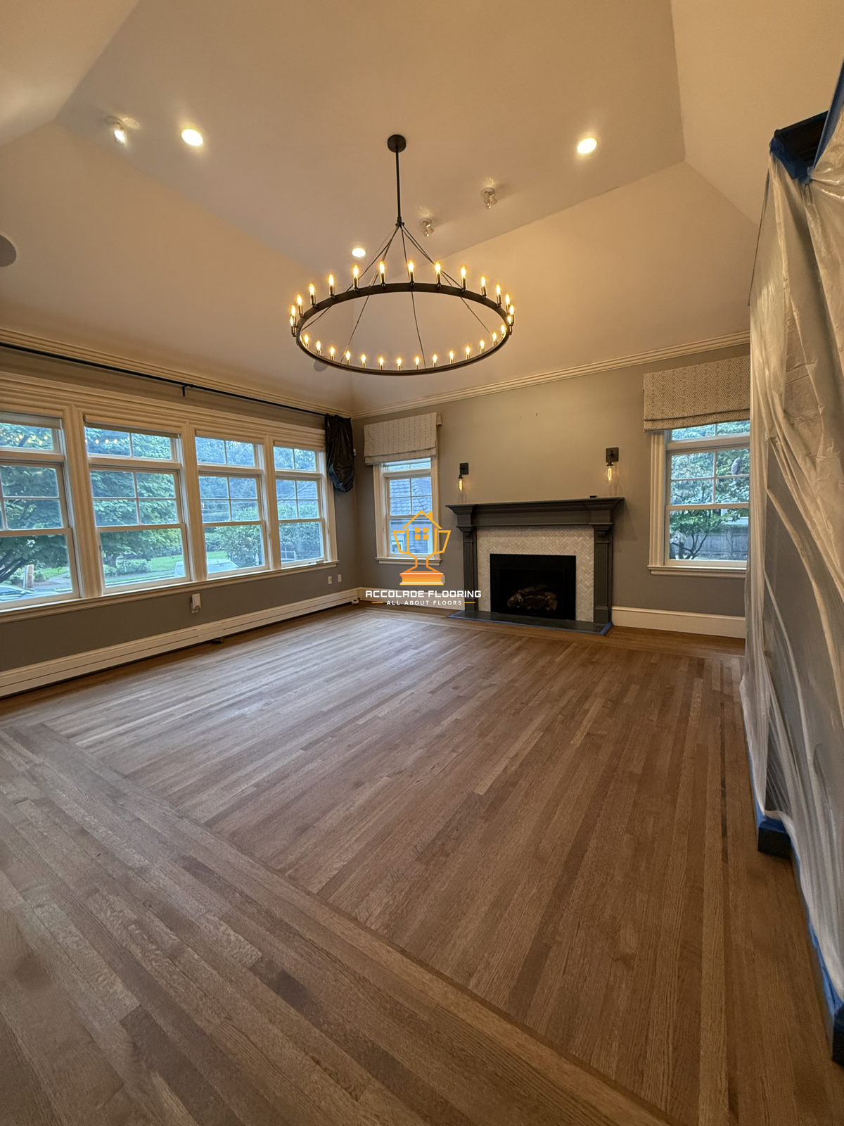 Living room with hardwood floors, large windows, a dark fireplace mantel, and a circular chandelier with lit candles.