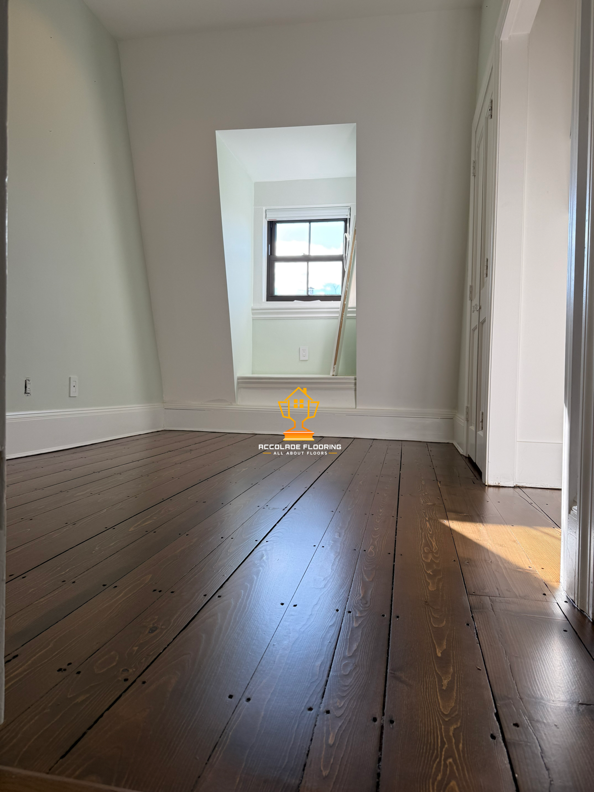 Empty room with wooden floor, white walls, a window, and a built-in closet. Logo for Accolade Flooring at the center of the floor.
