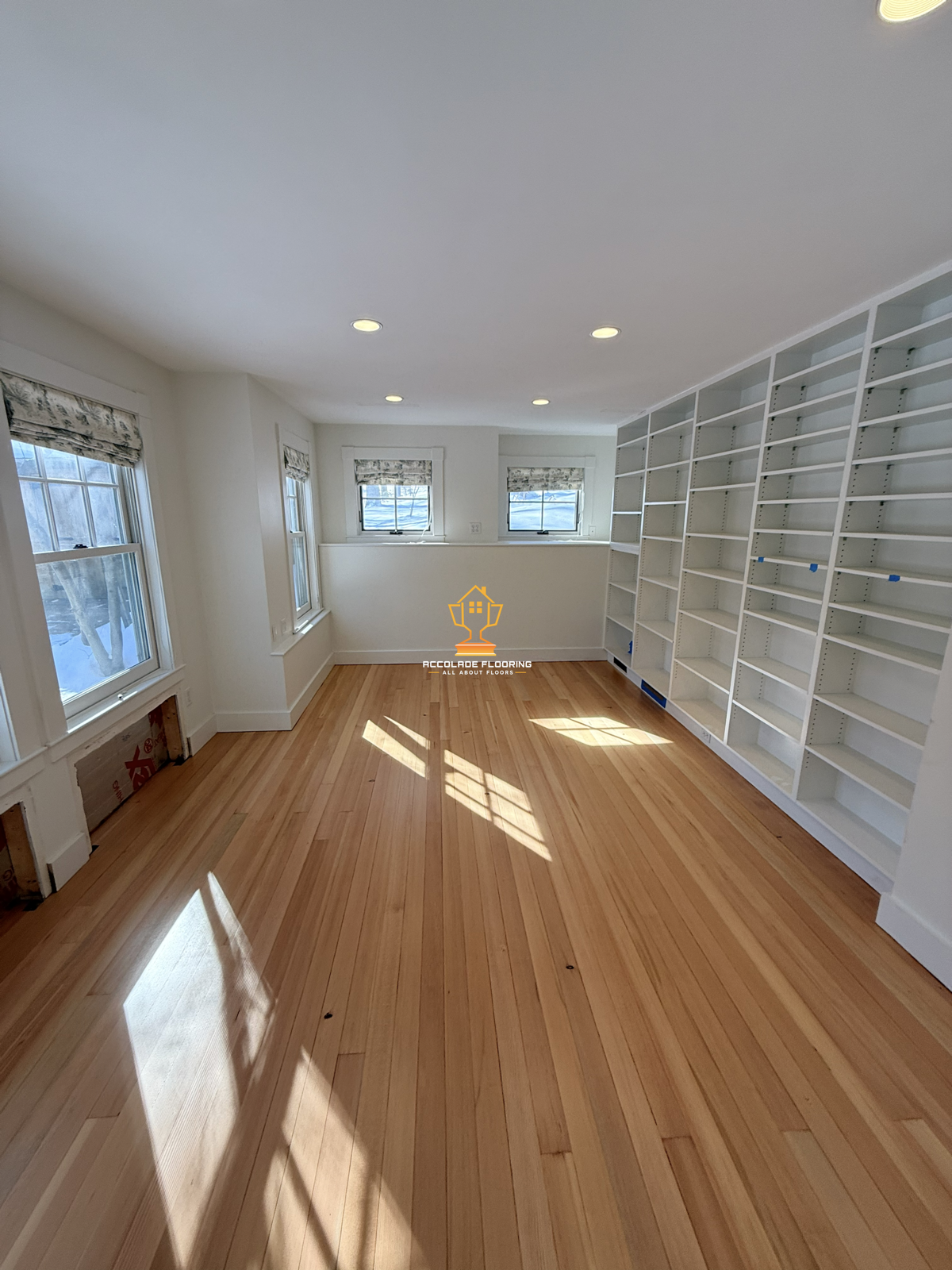 Room with hardwood flooring, multiple windows with blinds, white built-in shelves along one wall, and recessed ceiling lights.