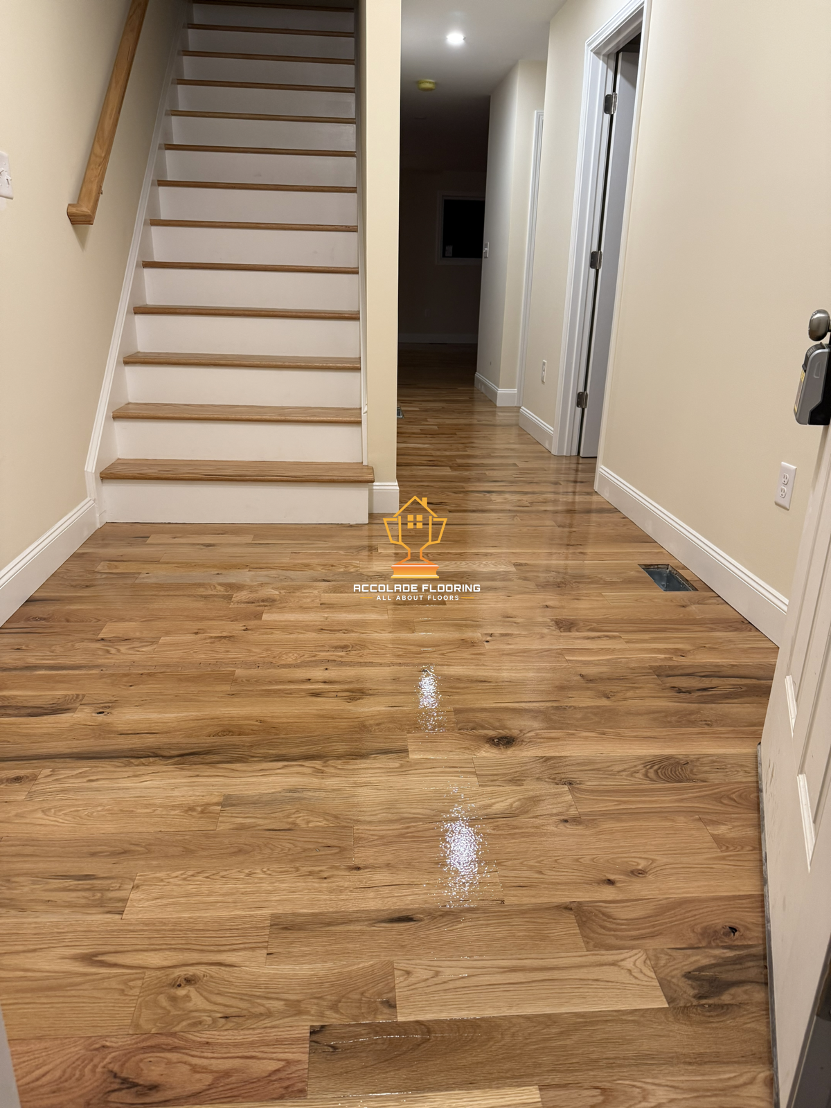 Interior view of a home with newly installed natural wood flooring, a staircase with wooden treads and white risers, and a hallway leading to other rooms. The floor is clean and shiny, reflecting light. The walls are painted a light color and there i