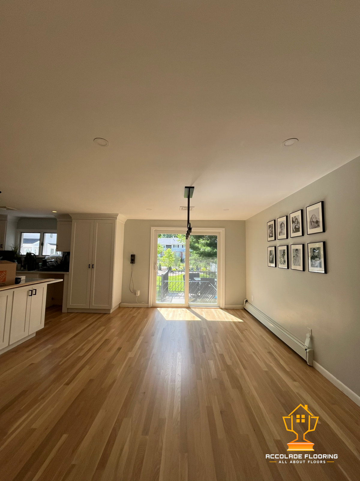 Empty living room with hardwood floors, a large sliding glass door leading to a balcony, framed photos on the wall, and kitchen area to the left.
