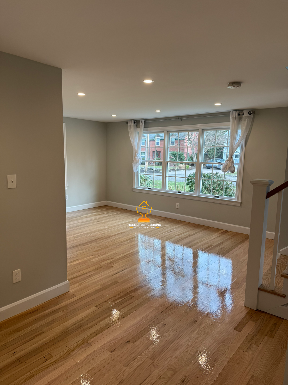 A bright living room with mandolin-colored hardwood floors, white walls, and large windows with sheer curtains. There are recessed ceiling lights and a staircase railing visible on the right.