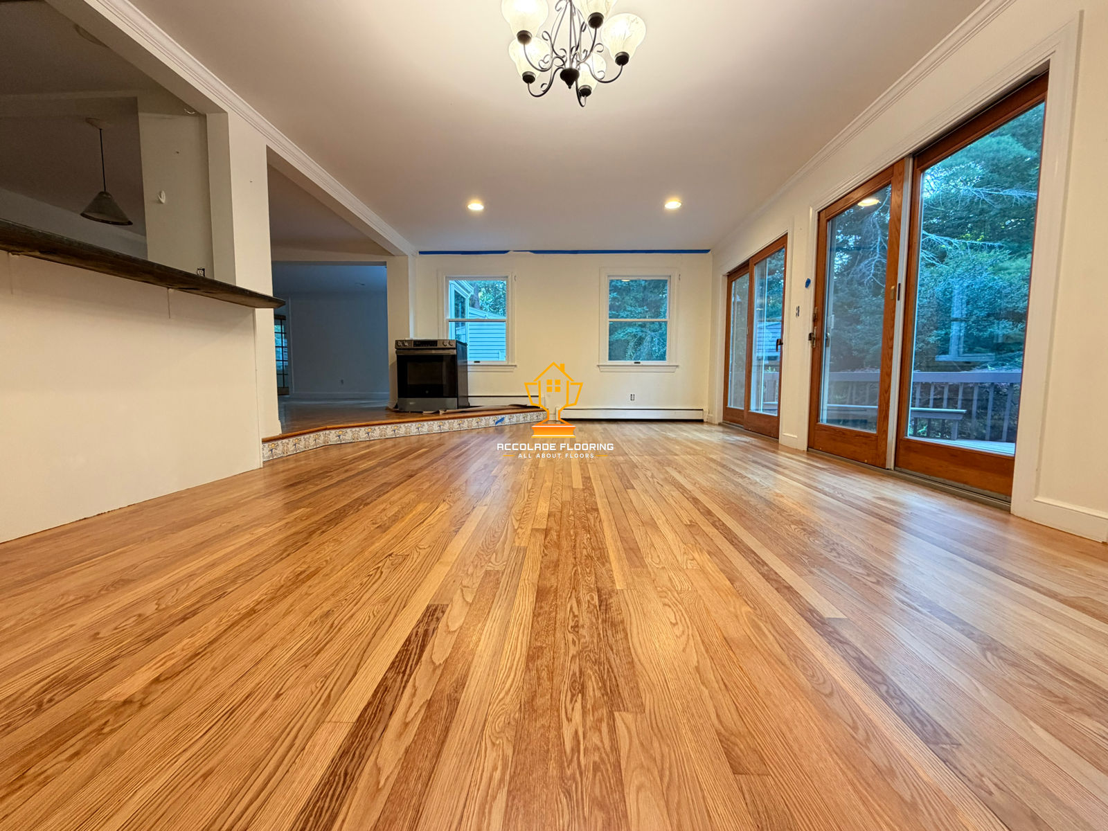 Living room with hardwood floor, large windows, and glass doors leading to a deck; chandelier hanging from ceiling; kitchen area with a stove in the background.