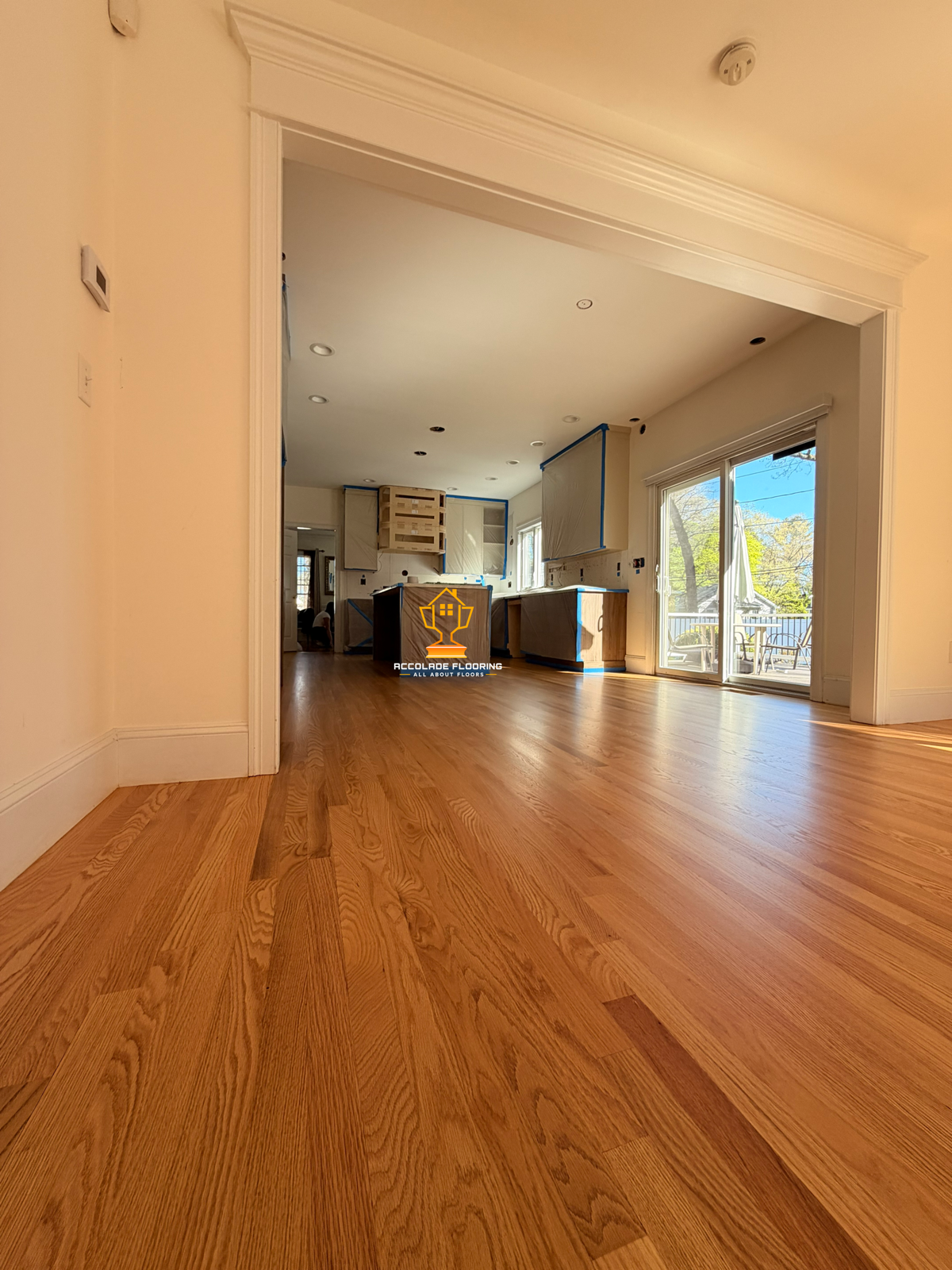A room with hardwood flooring, white walls, and large sliding glass doors leading to a patio. The room appears to be under renovation, with cabinets and counters covered in protective paper and tape. The ceiling has recessed lighting and a smoke dete