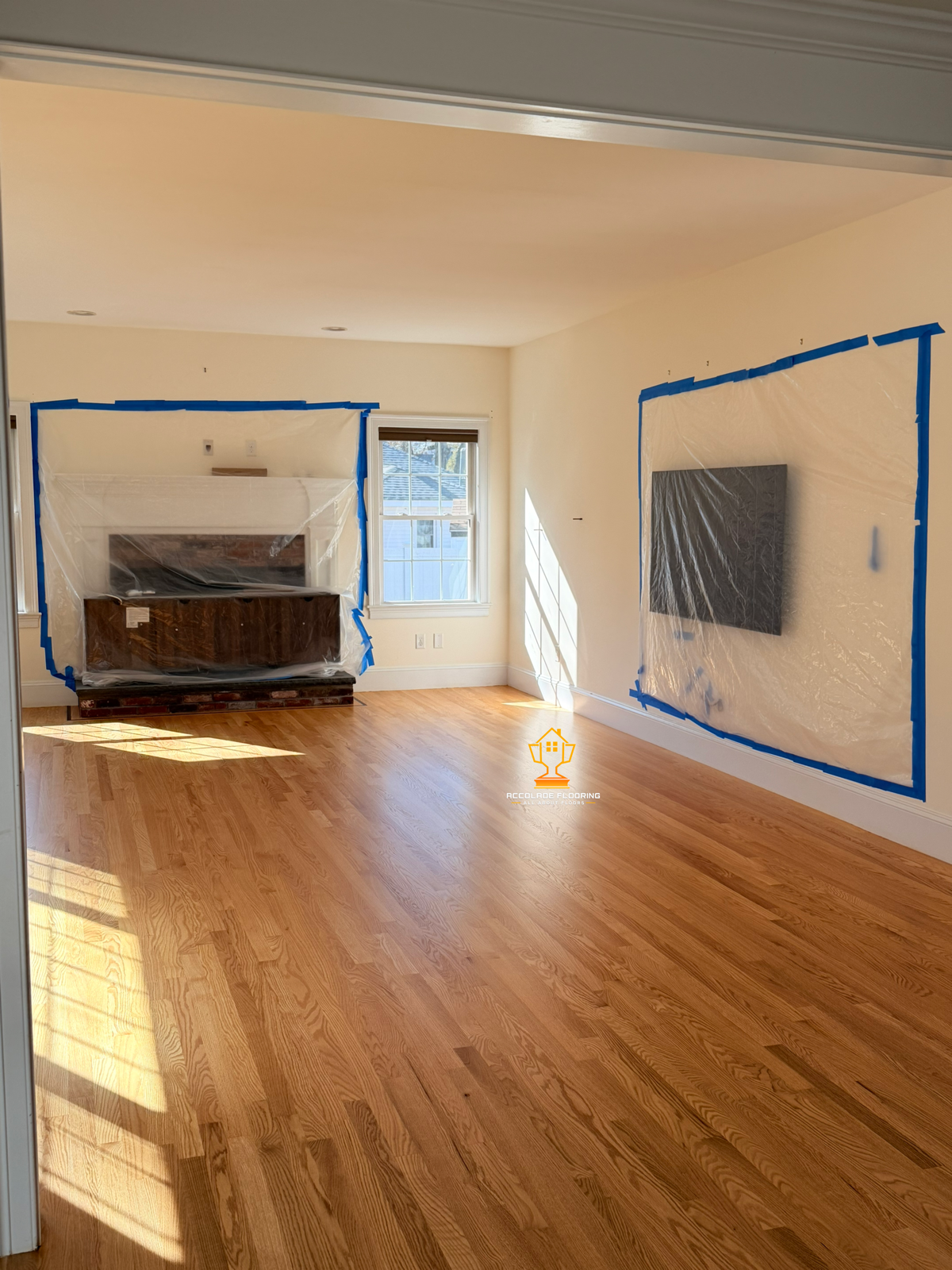 Living room with natural light, hardwood floor, wall-mounted TV covered with plastic, and a fireplace also covered with plastic, with blue painter's tape around the coverings for painting protection.